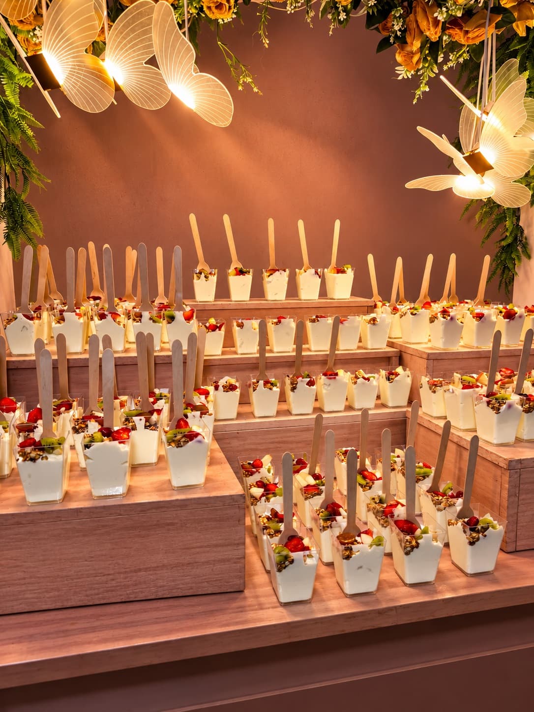 Rows of Norvilah parfait cups displayed on tiered wooden risers at an event, styled with hanging flowers and butterfly lights.